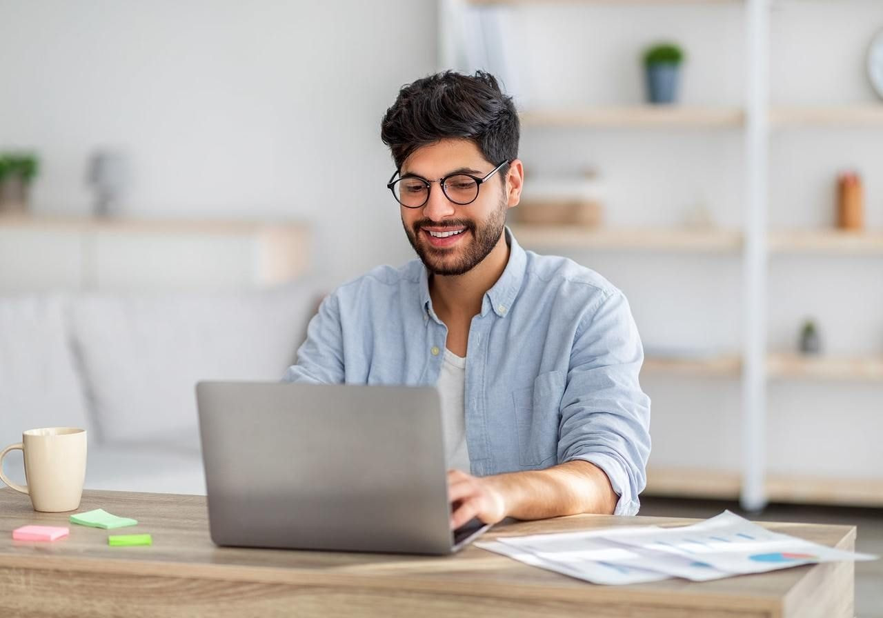 A young man in glasses is sitting at a desk and using a laptop to start a print-on-demand business